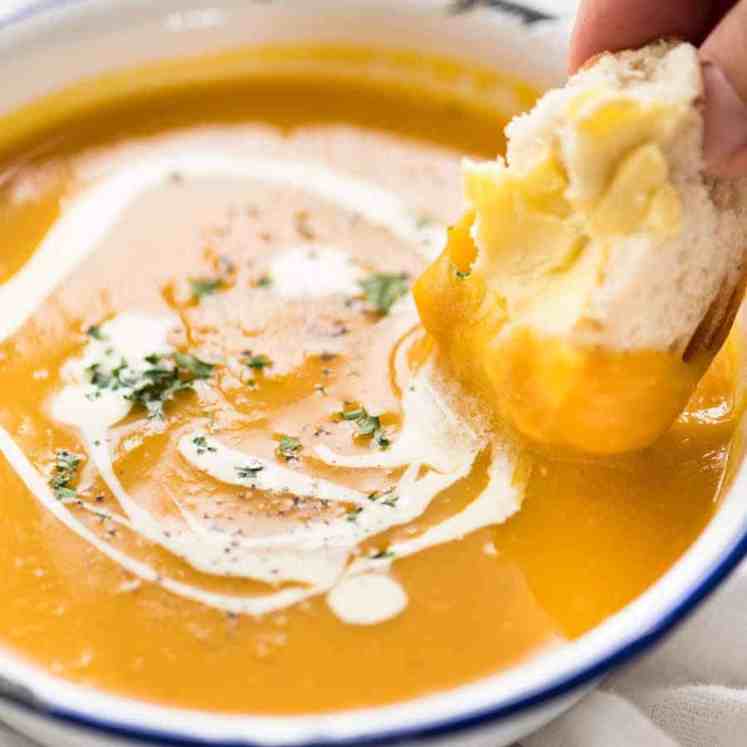 Close up of dunking crusty bread into thick and creamy pumpkin soup in a rustic white enamel bowl.